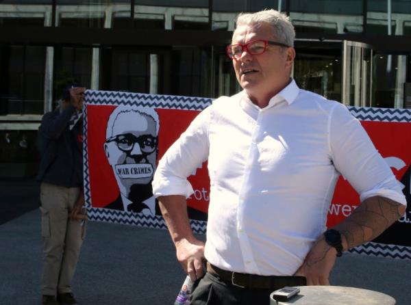 A man in a white shirt with red glasses stands in front of a red banner with an image of a man with his mouth covered with the words war crimes