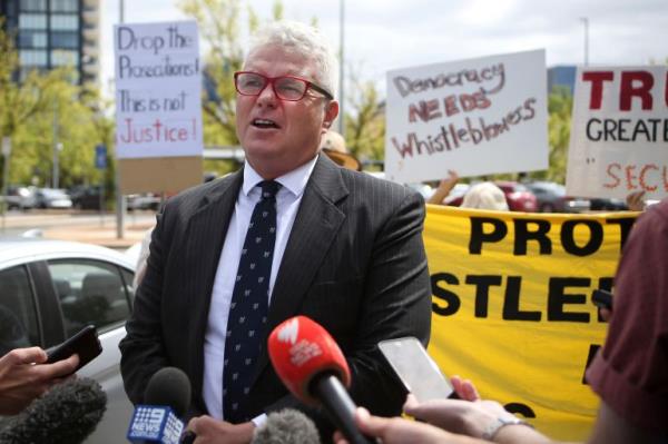 A man in a suit with several journalist micropho<em></em>nes in front of him stands in front of signs a<em></em>bout whistleblowers